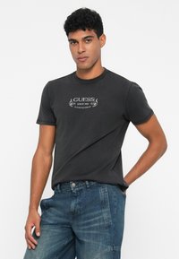 Joven con cabello rizado corto que lleva una camiseta negra con el logo "Guess" y jeans azules, de pie con una mano en el bolsillo.