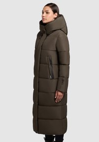 Garšs, olīvu zaļš pūkains mētelis ar augstu apkakli, rāvējslēdzēja kabatām un quiltējošu dizainu. Gluda tekstūra un kapuce pievieno siltumu.