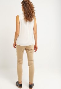 Top blanco sin mangas, pantalones ajustados beige y zapatos negros. Cabello rizado que cae hasta los hombros. Tejido liso, corte a medida, diseño minimalista.