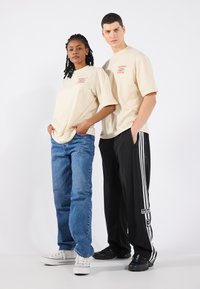 Beige t-skjorter med rødt trykk, blå jeans og svarte treningbukser med hvite striper. Fottøy inkluderer hvite joggesko og svarte sko.