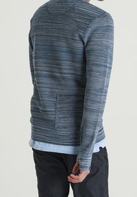 Niebieski sweter robiony na drutach z teksturowanym wzorem, okrągłym dekoltem i długimi rękawami. Zawiera kieszeń na plecach. Noszony na jasno-niebieskiej koszuli.