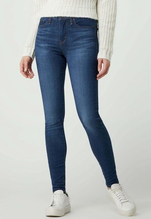 Kvinde iført mørkeblå skinny jeans, hvid strikket sweater og hvide sneakers mod en ensfarvet lys baggrund.