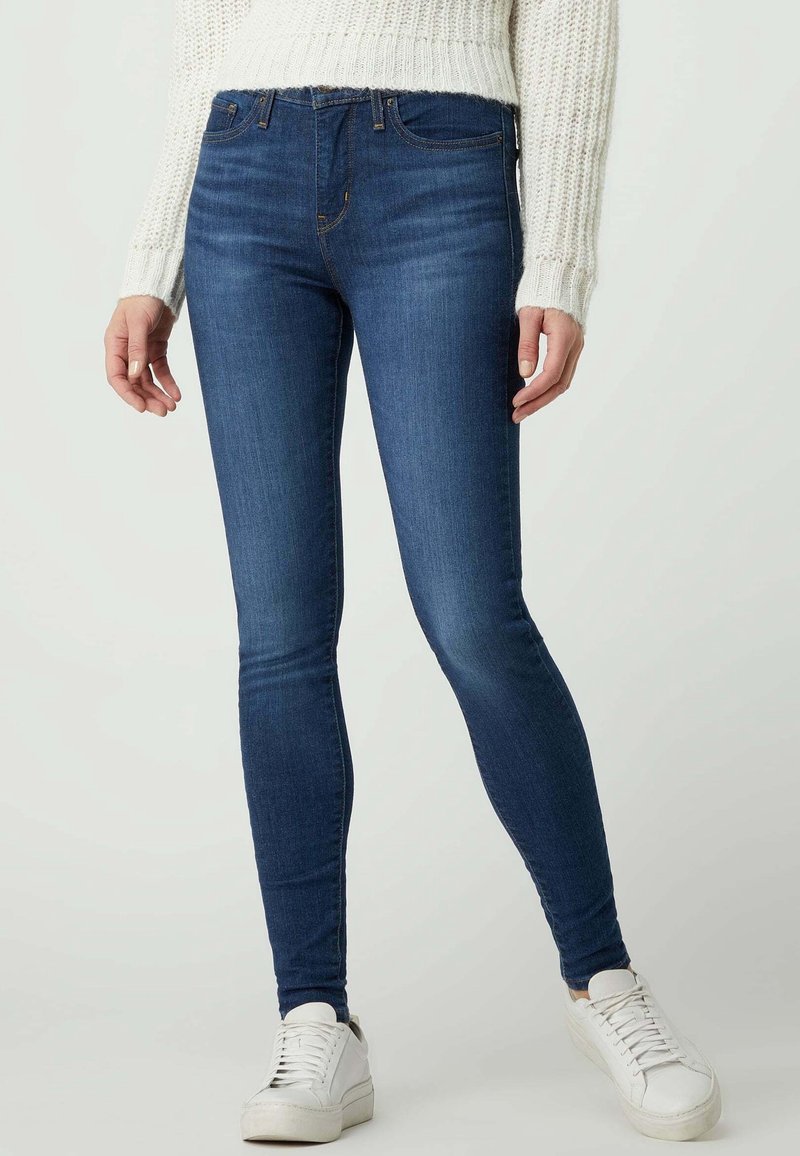 Femme portant un jean skinny bleu foncé, un pull en maille blanc et des baskets blanches sur un fond clair uni.