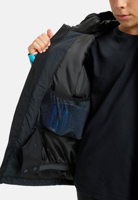 Schwarze Jacke mit glatter Außenseite, die eine Innentasche aus Mesh zeigt, in der blaue Gegenstände zu sehen sind. Enthält Druckknöpfe und Kordelzugverstellmöglichkeiten.