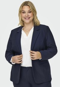Námořnický modrý blazer s klopami a dvěma předními kapsami, spárovaný s bílým topem. Hladká tkanina; model drží klopy blazeru.