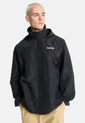 Mann mit kurzem blondem Haar, der eine schwarze Jacke mit Reißverschluss und Kapuze sowie beige Hose trägt, schaut seitlich vor weißem Hintergrund.