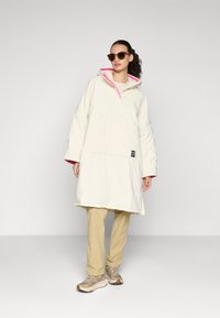 Nafršeni beli anorak z rožnatim notranjim podloženjem, ki ima pol-ziip in sprednjo žep. V kombinaciji z bež hlačami in svetlobruno športnimi copati.