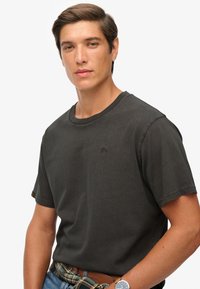 Czarnoszary bawełniany t-shirt z okrągłym dekoltem, krótkimi rękawami i subtelnym logo na piersi; zestawiony z plisowaną paskiem i dżinsami.