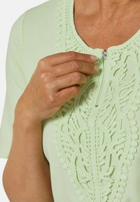 Blusa verde chiaro con un design in pizzo lungo il collo. Presenta una chiusura con zip sulla parte anteriore e maniche corte. Tessuto dalla texture morbida.