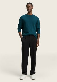 Türkis gestrickter Pullover mit Rundhalsausschnitt, kombiniert mit schwarzen, gefalteten Hosen und weißen Sneakers, vor einem neutralen Hintergrund präsentiert.