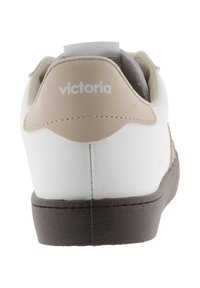 Baskets blanches avec un tirant beige à l'arrière et une semelle marron. Présente un matériau supérieur lisse et un logo embossé à l'arrière. Design et forme simples.