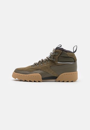Chaussure de randonnée montante vert olive avec semelle robuste couleur beige, œillets métalliques, languette de tirage et accents en texture suédée.