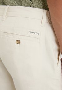 Light beige cotton pants featuring a back pocket with a button and a small white label reading "Marc O'Polo." Texture appears soft.