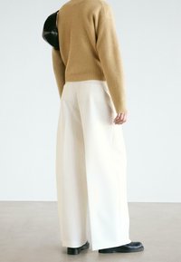 Beżowy prążkowany sweter z dzianiny w połączeniu z białymi, wysokim stanem i szerokimi nogawkami spodniami. Czarne skórzane buty dopełniają stylizację. Widok z tyłu.
