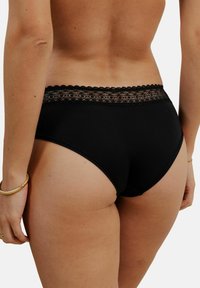 Culottes noires taille haute avec une ceinture en dentelle à bords festonnés et une texture de tissu lisse, présentées sous un angle latéral-arrière.