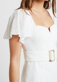 Biała sukienka z krótkimi, falbanistymi rękawami, teksturowanym materiałem z misternymi wzorami oraz szerokim pasem w talii z prostokątną klamrą.