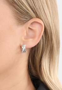Orecchini a cerchio in argento con un design intrecciato, caratterizzati da una superficie testurizzata e da dettagli trasparenti e scintillanti, che mettono in risalto una finitura metallica lucida.