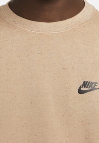 Close-up of a beige speckled Nike sweatshirt with a round neck and black Nike logo on the upper left chest area.