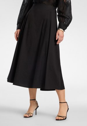 Frau trägt schwarzen Midi-Rock kombiniert mit durchsichtigem schwarzem Spitzenbluse mit Ärmeln aus Spitze und schwarzen, hochhackigen Sandalen mit Riemen, silbernes Armband am Handgelenk.