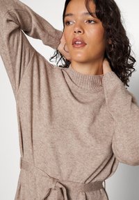Pull en maille beige clair avec un col côtelé, des manches longues et une taille ceinturée. Texture lisse et design simple.