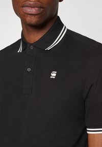 Černé polo tričko s strukturovaným límečkem s bílými pruhy. Má tříknopový zapínání a malý vyšívaný logo na hrudi.