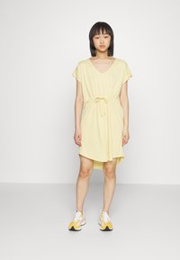 Vestido amarelo de manga curta com decote em V, cintura com cordão e um corte descontraído. Combinado com ténis de desporto brancos e amarelos.