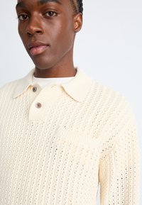 Suéter de punto en color crema, con un patrón texturizado, cuello con solapa, dos botones y un bolsillo en el pecho. Tejido suave y liviano.