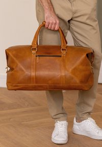 Persoon houdt grote bruin leren weekendtas met ritsvak vast, gekleed in beige broek en witte sneakers op een houten vloer.