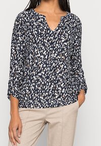 Navybluse mit einem weiß-braun gesprenkelten Muster. Sie verfügt über einen V-Ausschnitt, lange Ärmel mit umgeschlagenen Manschetten und einen weichen, strukturierten Stoff.