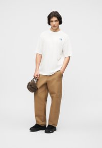 Νέος άνδρας που φοράει λευκό μπλουζάκι North Face, καφέ παντελόνι, μαύρα παπούτσια, κρατώντας μια μικρή καφέ τσάντα στο ένα χέρι, στέκεται μπροστά σε μονόχρωμο φόντο.