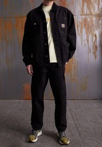Svart denimjacka med guldiga detaljer och fickor, kombinerad med matchande svarta byxor och olivgröna sneakers. Ljust färgad t-shirt under.