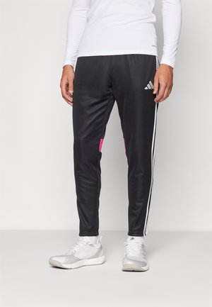 Personne portant un pantalon de survêtement Adidas noir avec des bandes blanches et des détails roses, des baskets blanches et un haut blanc à manches longues.