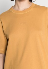 Mujer vistiendo una camiseta de cuello redondo amarilla mostaza con mangas cortas y un sutil logo de "Marc O'Polo" en el pecho.