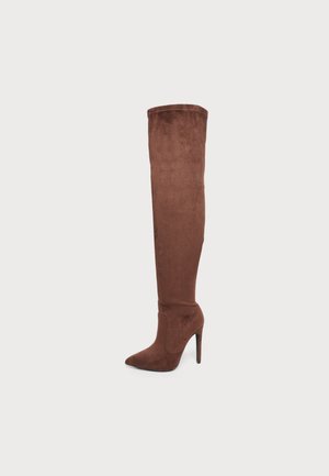 Botte stiletto en daim marron montant jusqu'à la cuisse, avec un bout pointu et un talon haut et fin sur un fond clair uni.