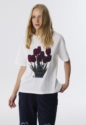 Camiseta blanca de algodón con un gráfico de tulipanes rojos oscuros y el texto "RÍE AMA VIVE REPITE". Corte relajado con mangas cortas.