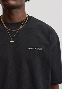Person iført en sort Pegador T-shirt med to guldkæder, den ene med et korsvedhæng, mod en ensartet lys baggrund.