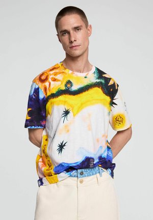 Mann mit kurzen Haaren, der ein kurzärmeliges abstraktes mehrfarbiges Hemd und hellbeige Hose vor einem einfarbigen Hintergrund trägt.