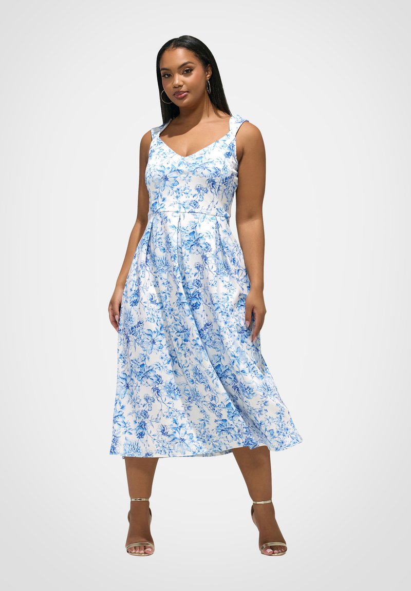 Frau trägt ein ärmelloses, weißes Midikleid mit blauem Blumenmuster und goldenen Knöchelriemen-High Heels, steht vor einem schlichten Hintergrund.