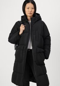 Mustat puhvitakki, jossa on huppu, tilava peitetty muotoilu, suuret etutaskut ja puolipitkä leikkaus. Valmistettu synteettisestä materiaalista.