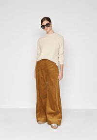 Beiger Pullover aus weichem Stoff, kombiniert mit weit geschnittenen, gerippten, senfgelben Cordhosen und beigen Slippern. Schwarze übergroße Sonnenbrille.