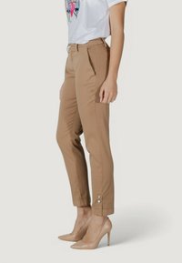 Pantalon beige sur mesure avec poches latérales, ourlets retroussés avec boutons en accent, associé à des chaussures à talons beiges et un t-shirt blanc avec un motif graphique.
