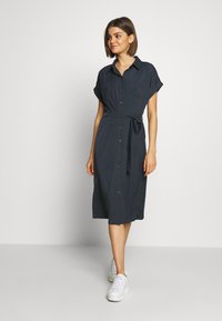 Vestito camicia blu navy, lungo fino al ginocchio, con maniche corte arrotolate, bottoni sulla parte anteriore, cintura in vita e una singola tasca sul petto.