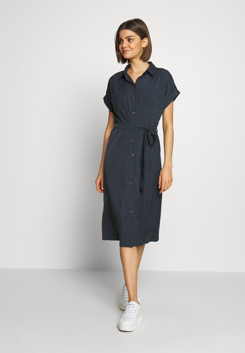 Vestito camicia blu navy, lungo fino al ginocchio, con maniche corte arrotolate, bottoni sulla parte anteriore, cintura in vita e una singola tasca sul petto.