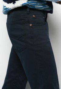 Gros plan sur une personne portant un pantalon bleu marine foncé avec des rivets en cuivre et un pull multicolore à motifs rentré.
