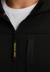 Sudadera con cremallera negra que presenta una tela suave y texturizada, una etiqueta de logo amarilla en contraste y una cremallera resistente. Debajo hay una camiseta blanca.
