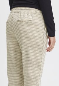Elastic-waist trousers in a light beige material, featuring a small brown check pattern and a back pocket. Textured surface and relaxed fit.
