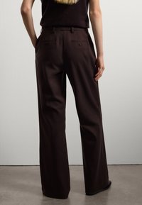 Persoon in hoge taille, wijde donkerbruine broek met achterste knoopzakken, staande tegen een effen lichte muur.