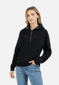 Felpa nera con zip, con coulisse, maniche raglan e una texture morbida. Abbinata a jeans azzurri chiari. Design minimalista.