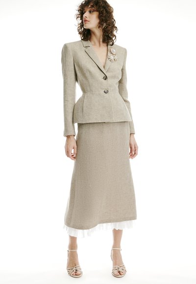 Mujer con blazer beige a medida con un broche decorativo y falda larga a juego sobre un dobladillo blanco con volantes, combinada con sandalias de tiras.