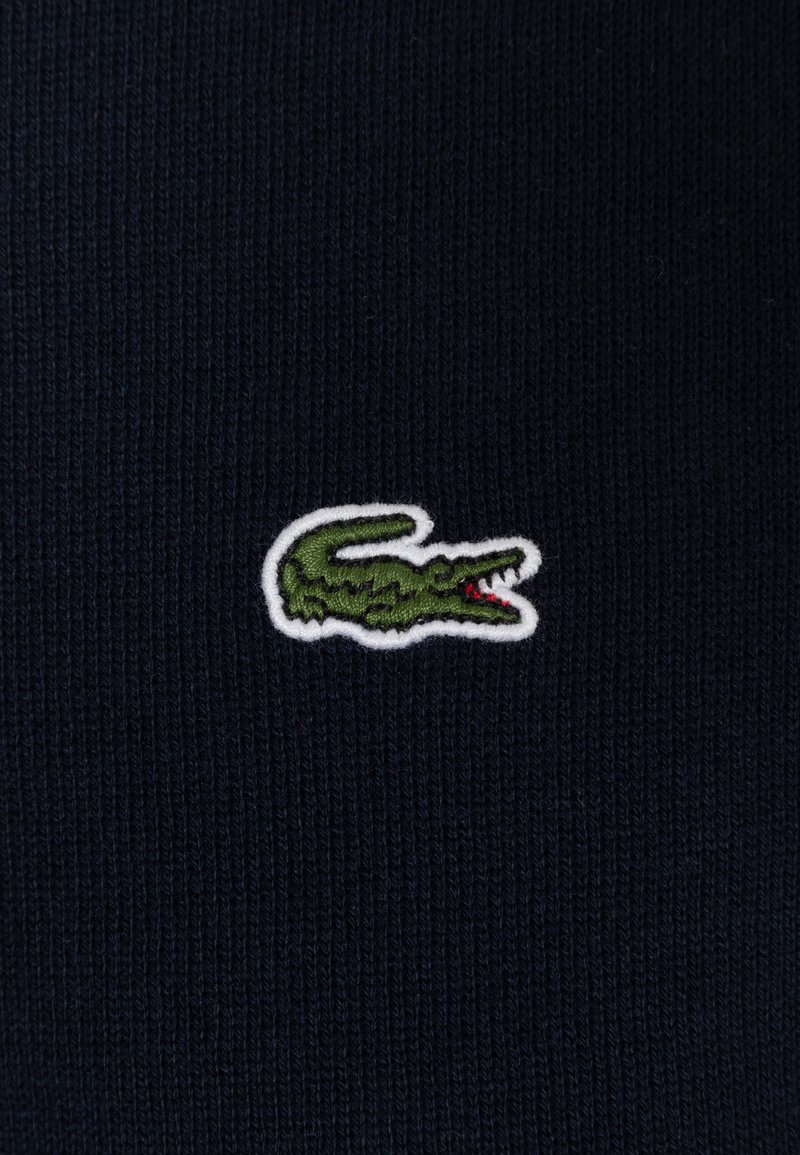 Il tessuto a maglia blu navy presenta un logo di coccodrillo verde con dettagli rossi, bordato da ricami bianchi. La texture è rigata e liscia.